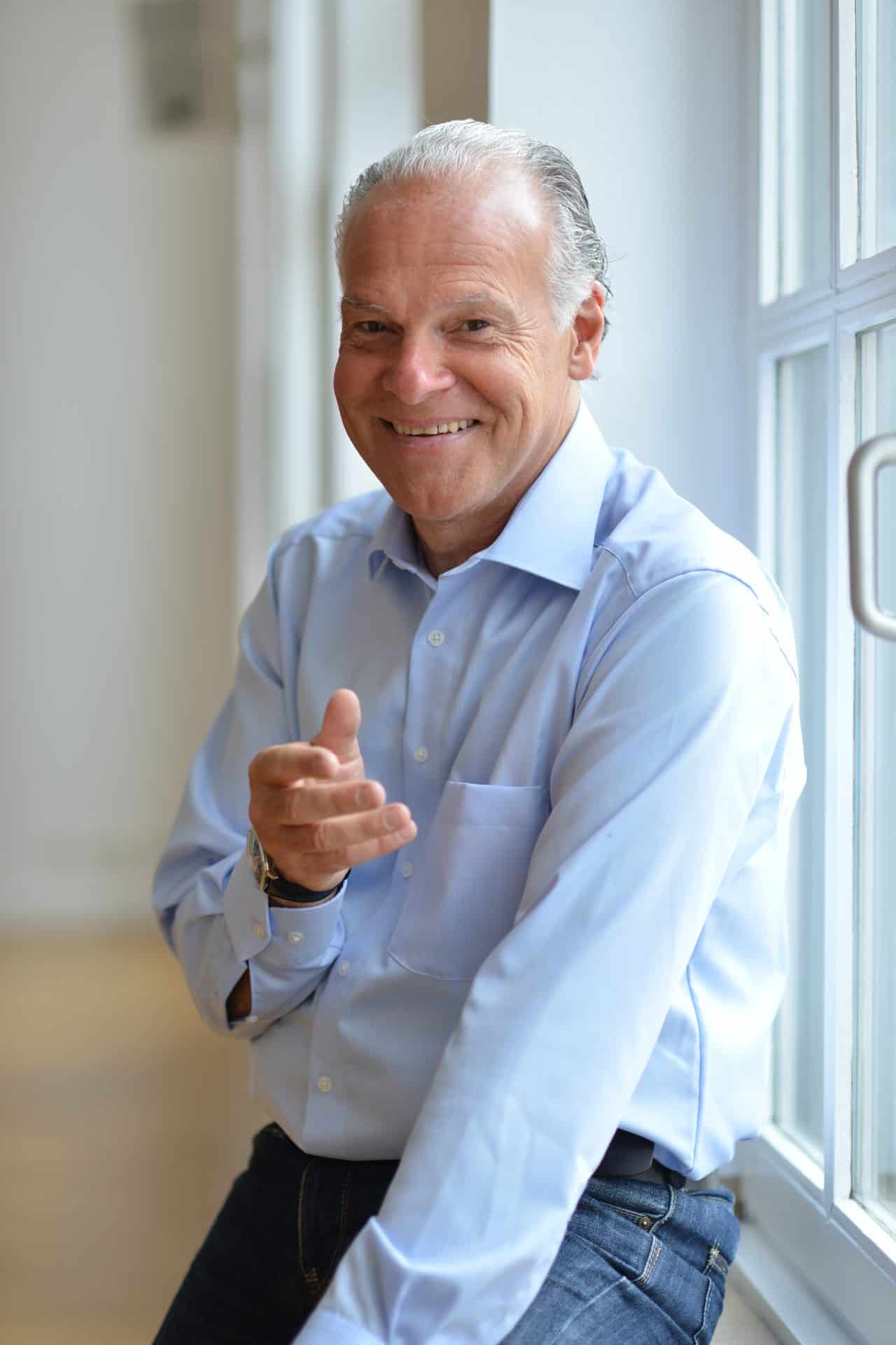Rolf Schmitz-Malburg lächelt und zeigt auf die Kamera – Porträt im Hochformat